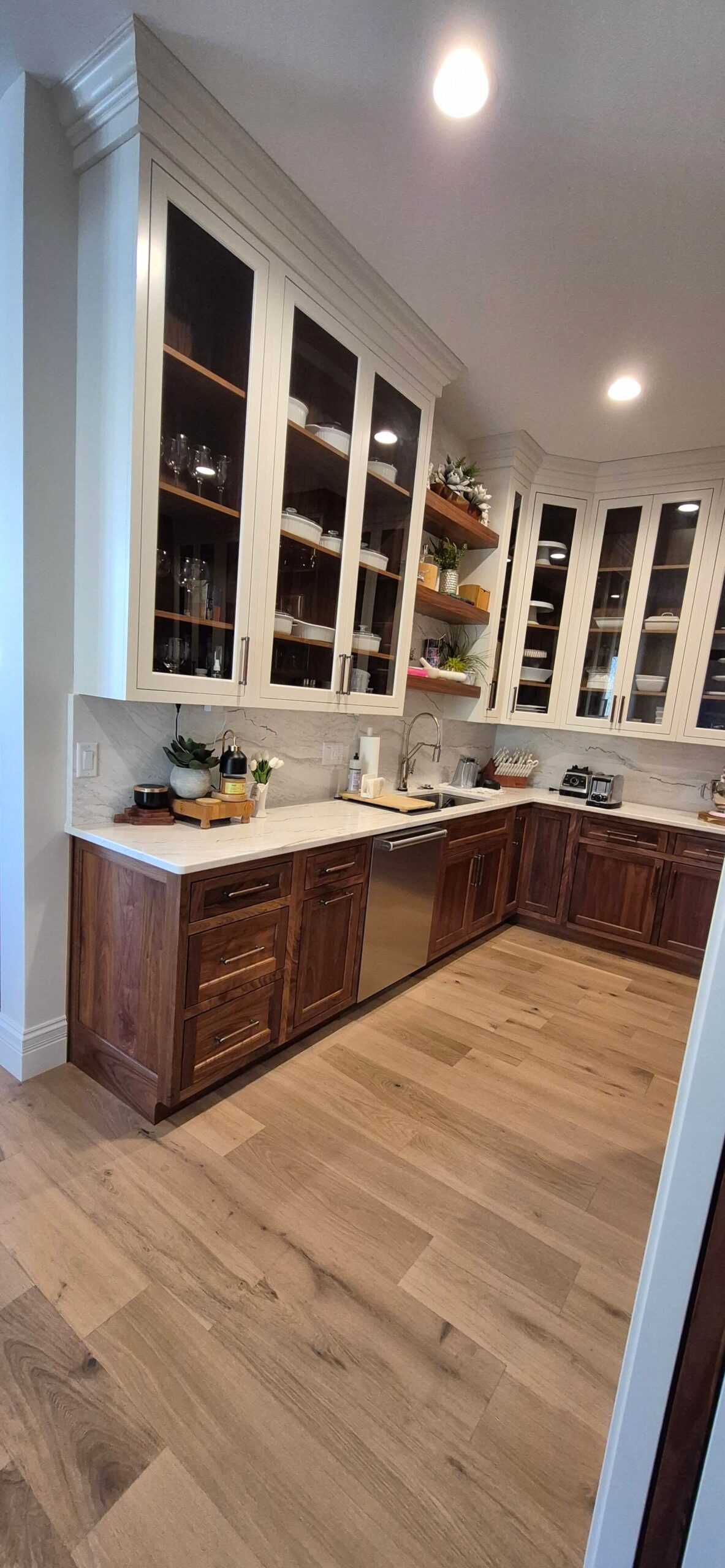 Creative Cabinetry Bloomfield Iowa Modern kitchen with wooden lower cabinets, white upper cabinets with glass doors, light wood flooring, and built-in appliances under bright ceiling lights.