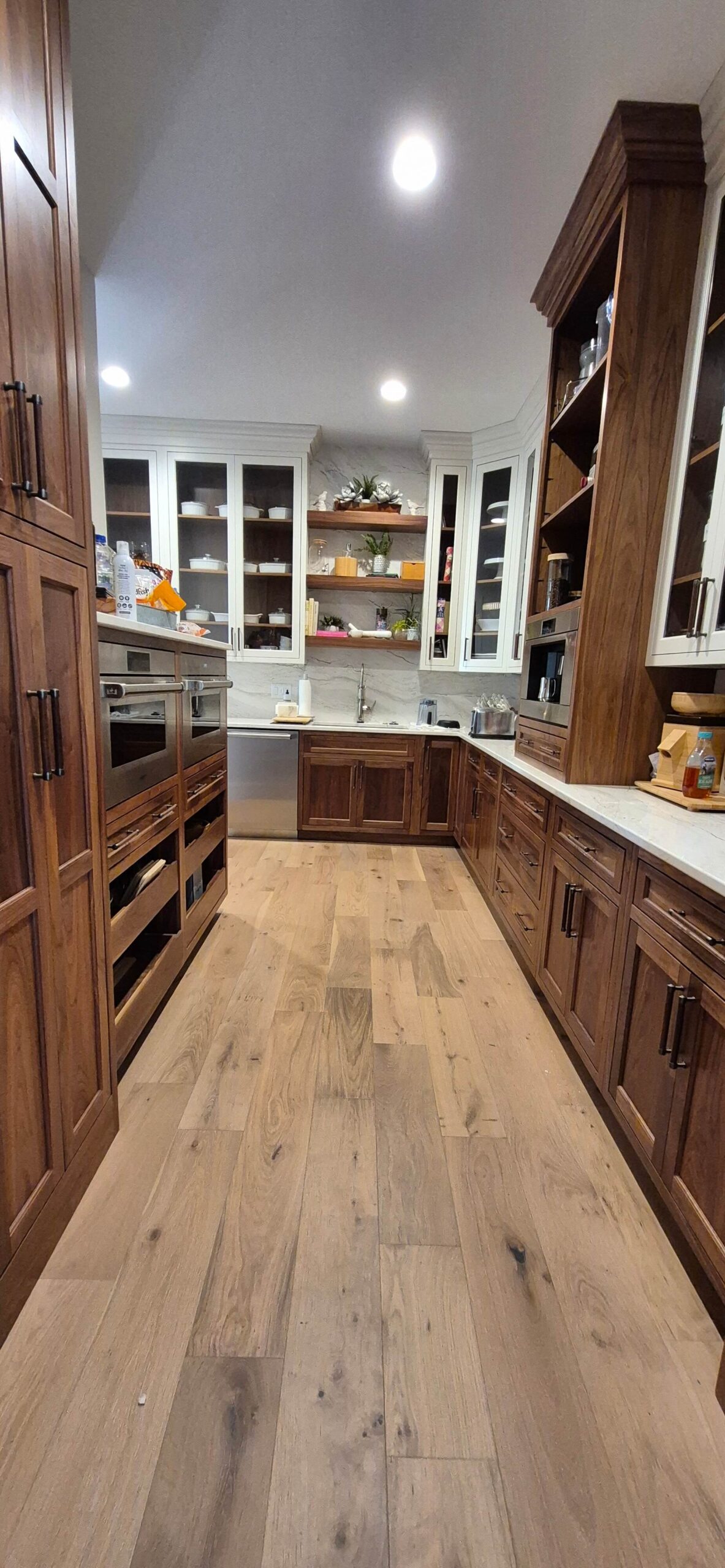 Creative Cabinetry Bloomfield Iowa Modern kitchen with wooden cabinets, light wood flooring, white countertops, stainless steel appliances, and open shelves displaying dishes and kitchen items.