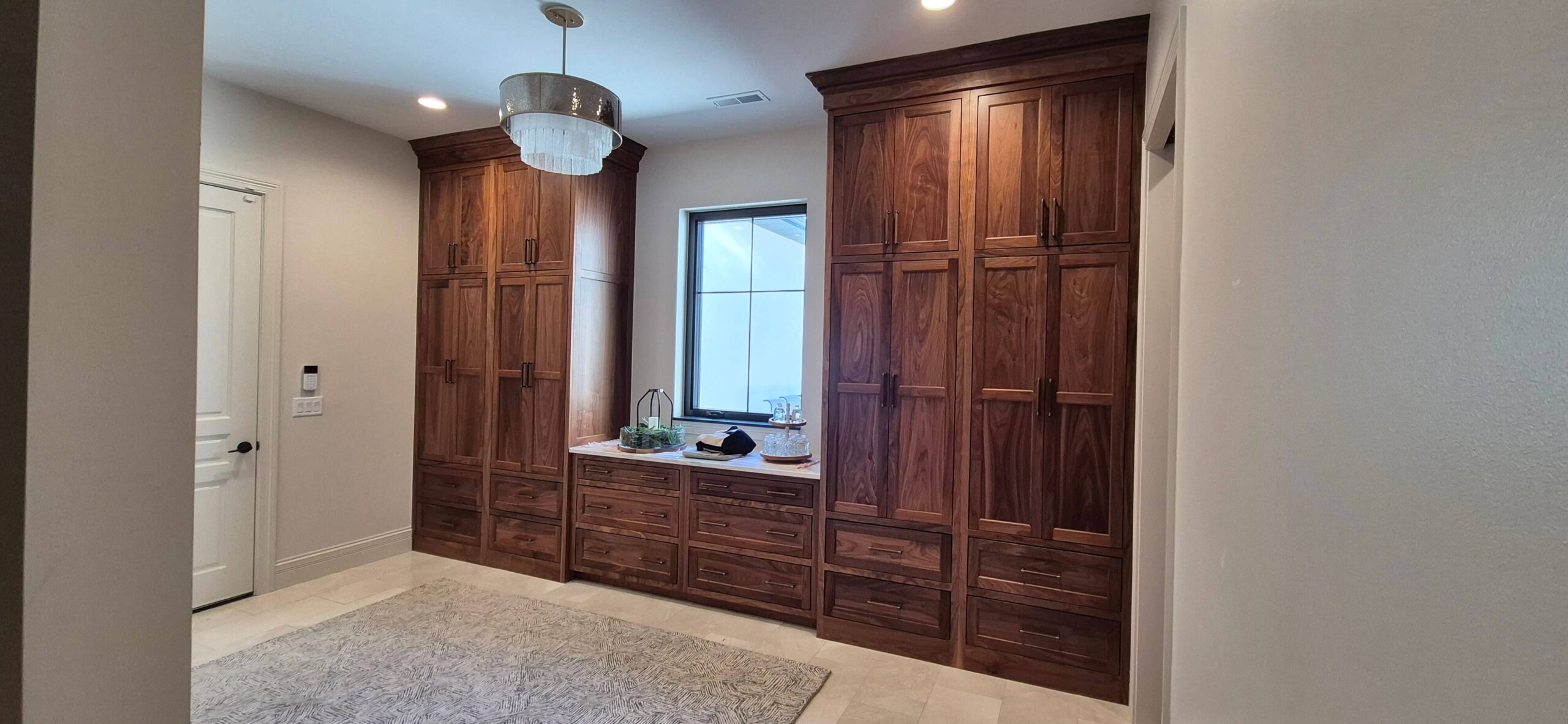Creative Cabinetry Bloomfield Iowa Spacious room with large wooden cabinets, a built-in countertop with decor items, a window in the center, and a modern chandelier above a light-colored rug.