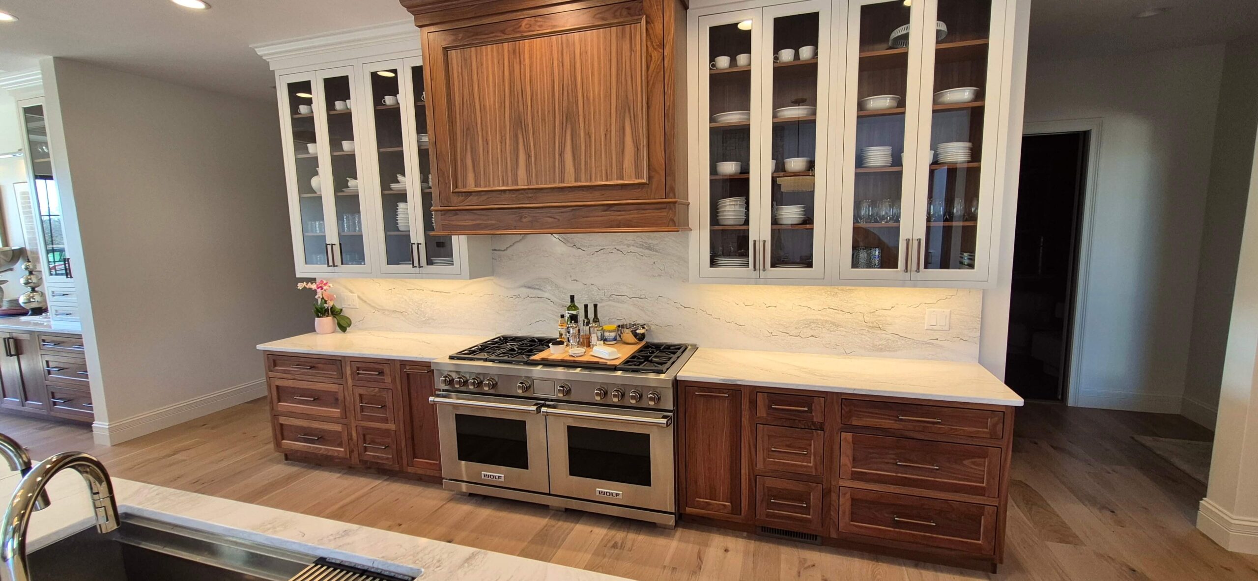 Creative Cabinetry Bloomfield Iowa Modern kitchen with stainless steel stove and oven, wooden cabinets, glass-front upper cabinets displaying dishes, and a marble backsplash.