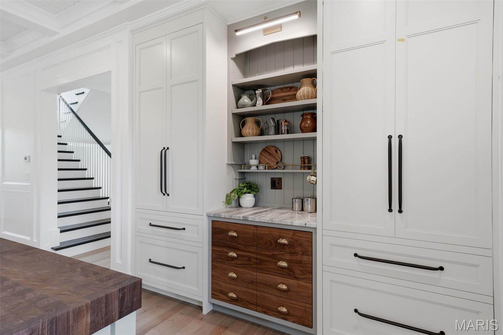 Kenrose Kitchens Custom Cabinetry Arthur IL A built-in kitchen cabinet with open shelves displays various dishes and decor items, surrounded by white cabinetry and a wood countertop. A staircase is visible in the background.