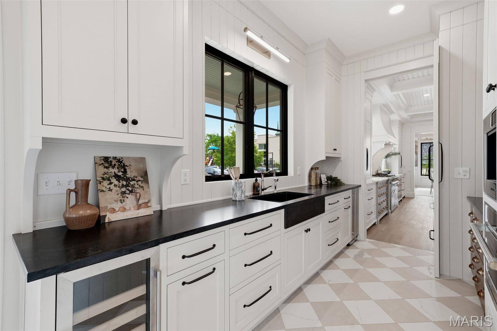 Kenrose Kitchens Custom Cabinetry Arthur IL A modern kitchen with white cabinets, black countertops, a window over the sink, built-in appliances, and checkered tile floor. A hallway leads to another kitchen area in the background.