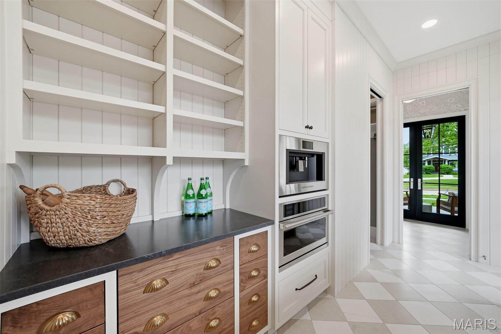 Kenrose Kitchens Custom Cabinetry Arthur IL Modern kitchen pantry with empty white shelves, wood drawers, built-in ovens, a basket, and three glass water bottles on a black countertop. Doorway leads to a bright room with large windows.