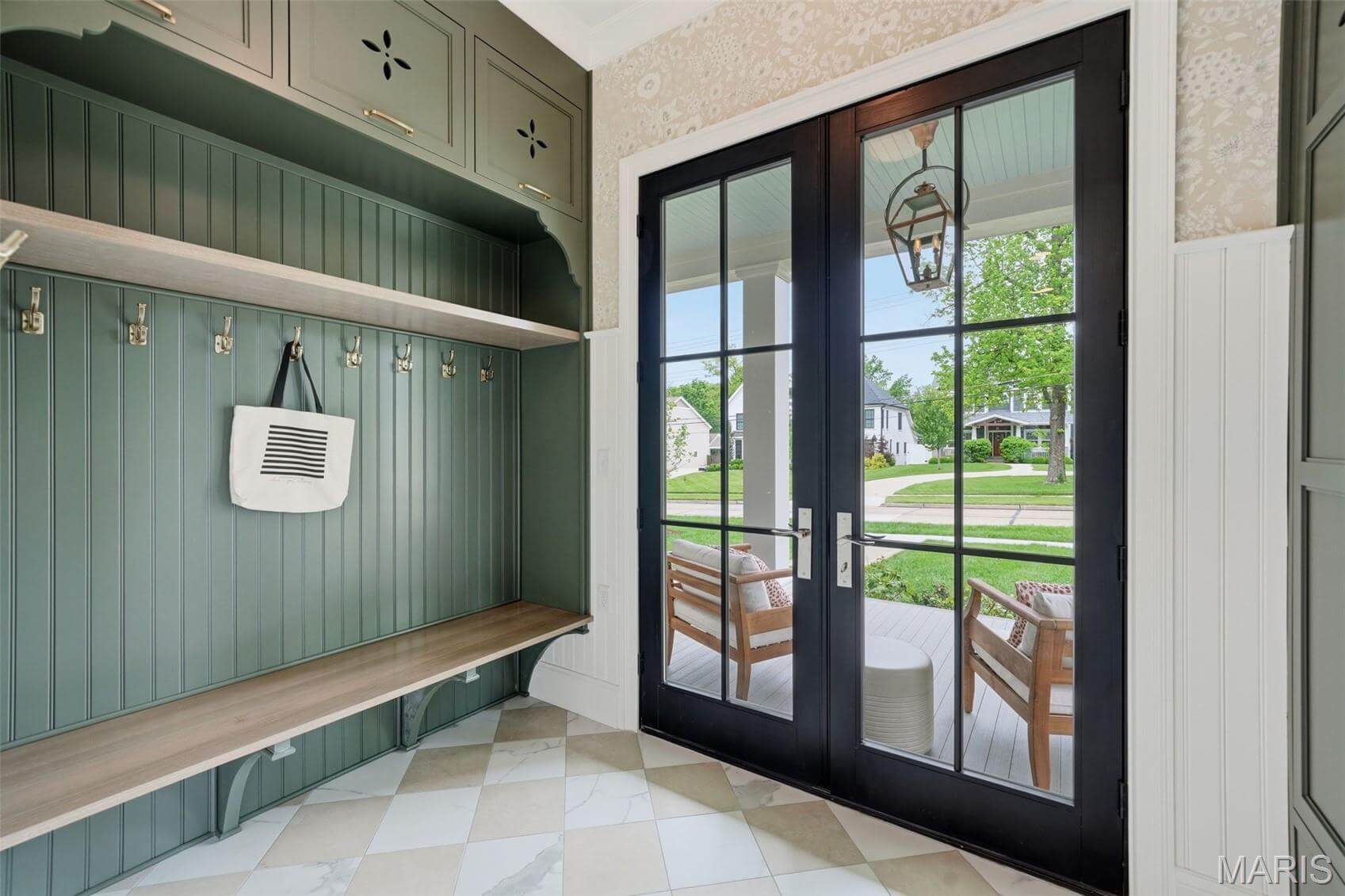 Kenrose Kitchens Custom Cabinetry Arthur IL A mudroom with green built-in storage, hooks, a bench, checkered tile floor, and glass double doors leading to a porch with two chairs and a view of houses outside.