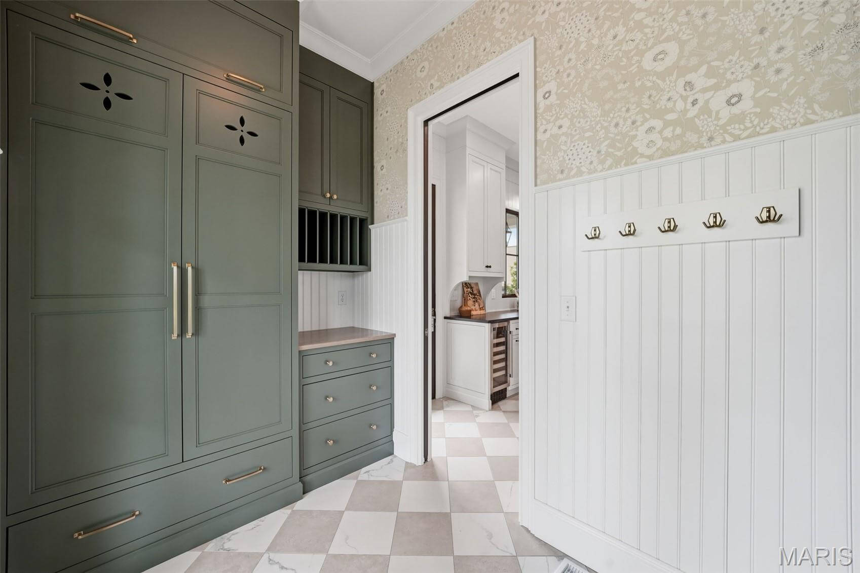 Kenrose Kitchens Custom Cabinetry Arthur IL Green built-in cabinets with gold handles, checkered tile floor, floral wallpaper, white wainscoting, and wall hooks in a transitional hallway leading to a kitchen.