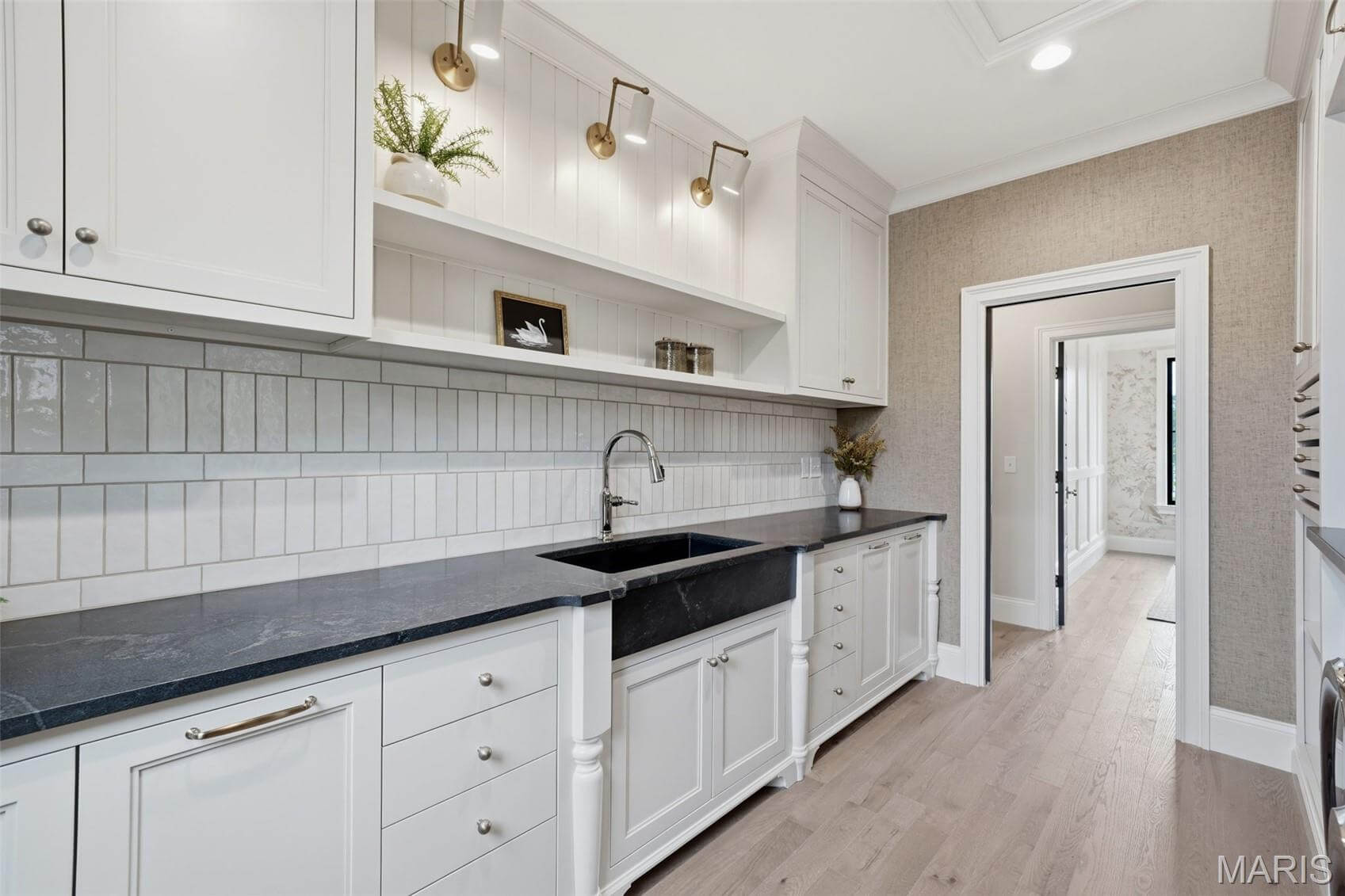Kenrose Kitchens Custom Cabinetry Arthur IL Modern kitchen with white cabinets, black countertops, a black farmhouse sink, wall-mounted lights, open shelving, and light wood flooring. A hallway is visible in the background.
