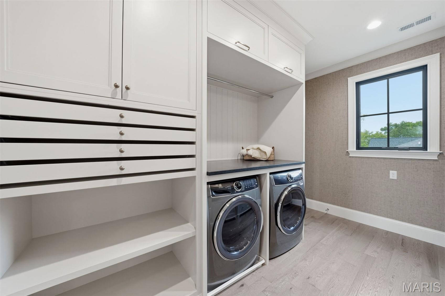 Kenrose Kitchens Custom Cabinetry Arthur IL Modern laundry room with front-loading washer and dryer, white built-in cabinets, open shelves, and a window letting in natural light.