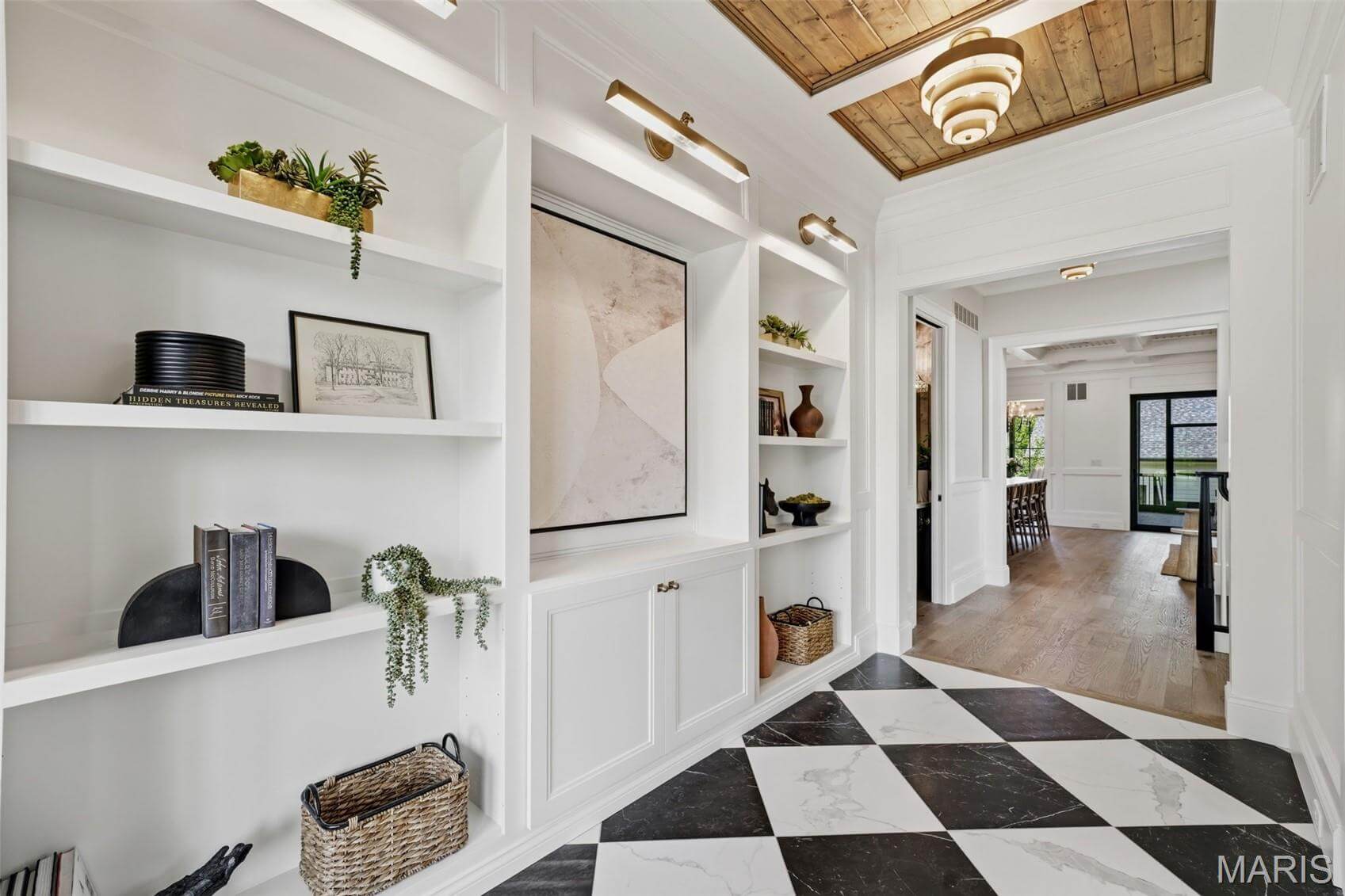 Kenrose Kitchens Custom Cabinetry Arthur IL A hallway with black-and-white checkered floor, built-in white shelves with decor, a wooden ceiling inset, and a view into a dining area with large windows.