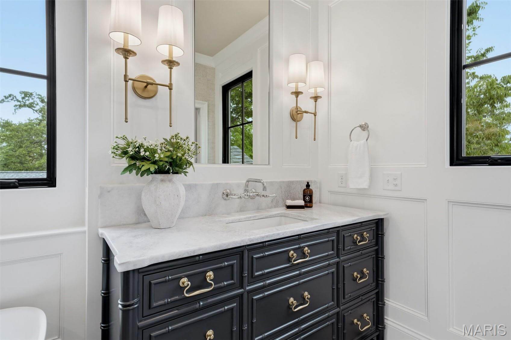 Kenrose Kitchens Custom Cabinetry Arthur IL A bathroom vanity with a white marble countertop, black cabinets, silver fixtures, wall-mounted lights, a vase with greenery, and a hand towel by a window.
