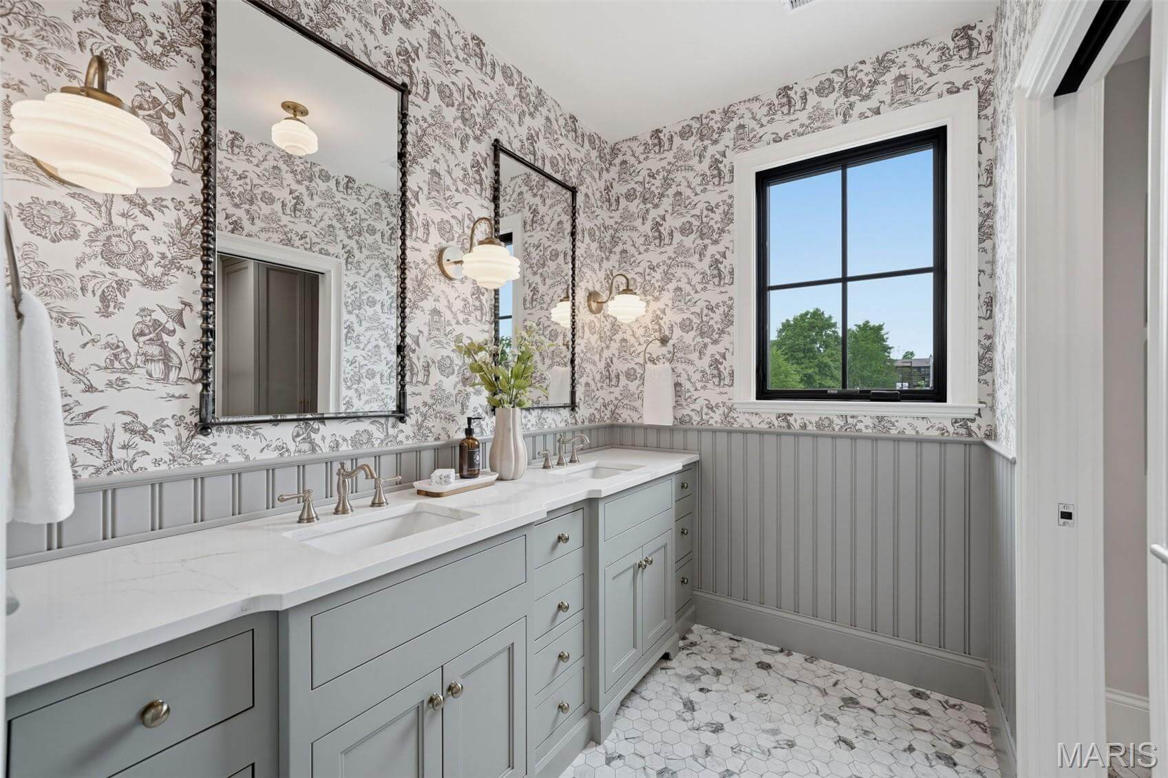 Kenrose Kitchens Custom Cabinetry Arthur IL Bathroom with double sinks, gray cabinetry, floral wallpaper, two mirrors, wall sconces, a window, decorative vase, and hexagonal tile flooring.
