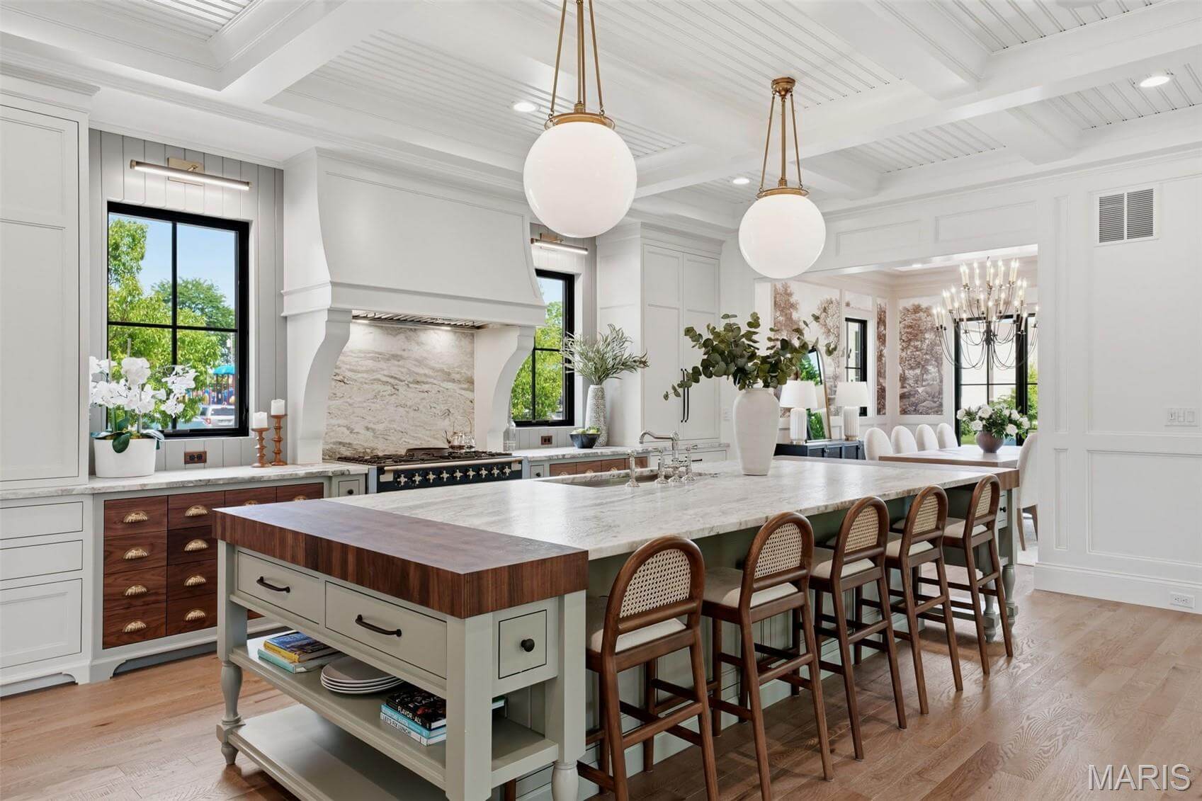 Kenrose Kitchens Custom Cabinetry Arthur IL Spacious kitchen with a large marble island, wooden accents, four chairs, modern pendant lights, and white cabinetry, open to a dining area with a chandelier.