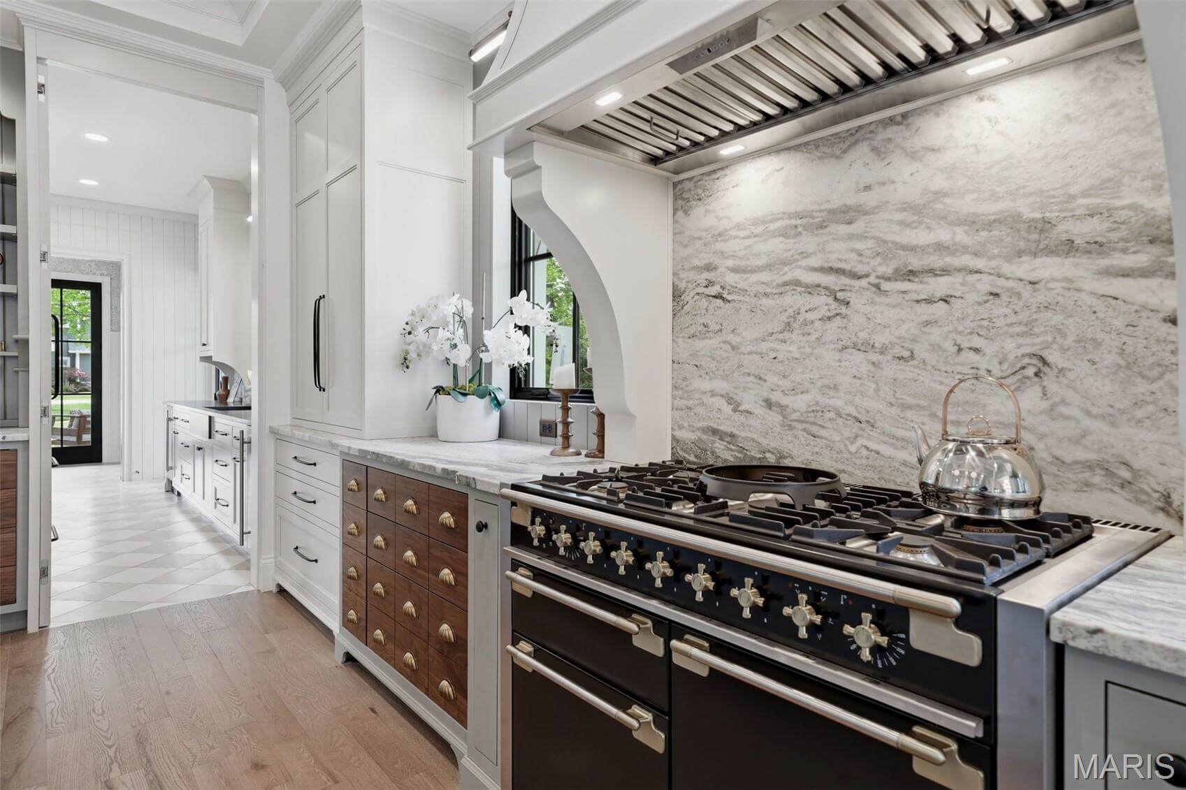 Kenrose Kitchens Custom Cabinetry Arthur IL A modern kitchen with a large black and stainless steel stove, a marble backsplash, white cabinetry, and wooden floors. A kettle sits on the stove and an orchid is on the counter.