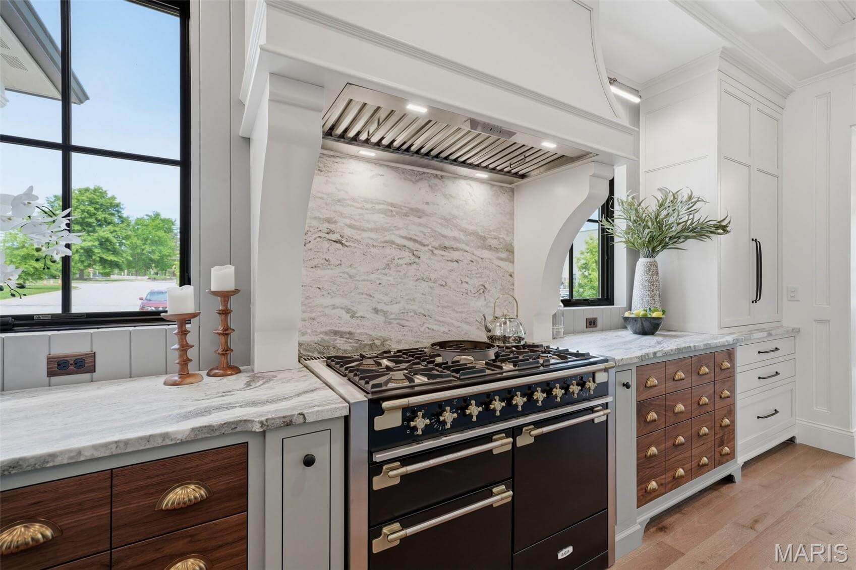 Kenrose Kitchens Custom Cabinetry Arthur IL Modern kitchen with a black and brass stove, marble backsplash, white cabinetry, and large windows providing natural light.