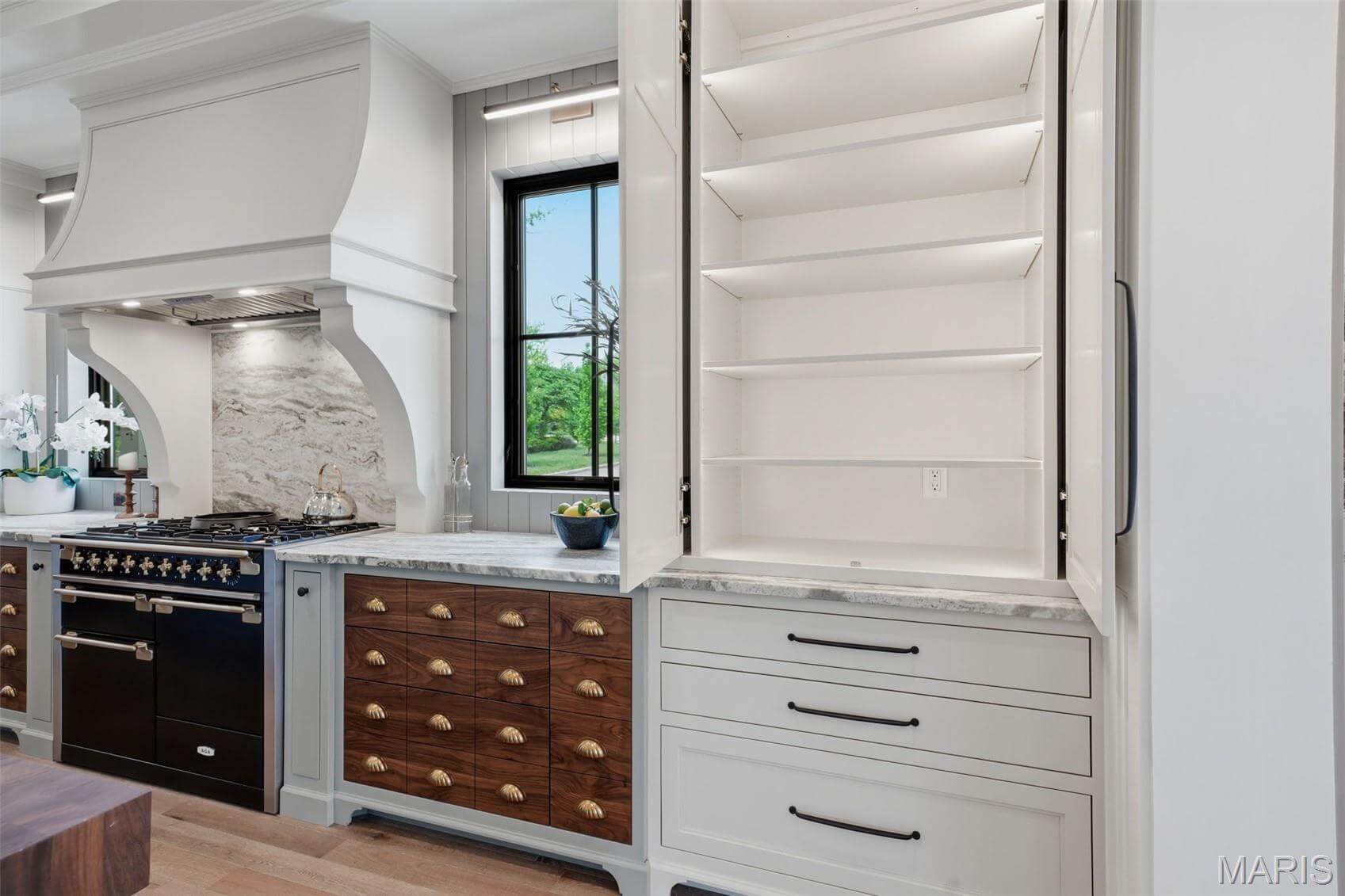 Kenrose Kitchens Custom Cabinetry Arthur IL Modern kitchen with a black stove, marble backsplash, wooden drawers, and an open pantry cabinet with white shelves and a marble countertop.