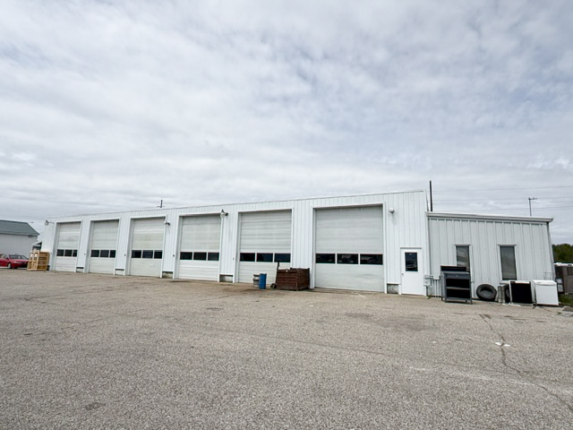 A large white industrial building with six garage doors and a small side office, set on a paved lot under a cloudy sky.