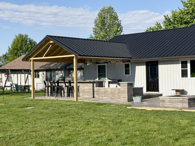 A backyard patio with a wooden pergola, outdoor bar seating, and a small built-in bench beside a house with a dark metal roof. Grass lawn and trees in the background.
