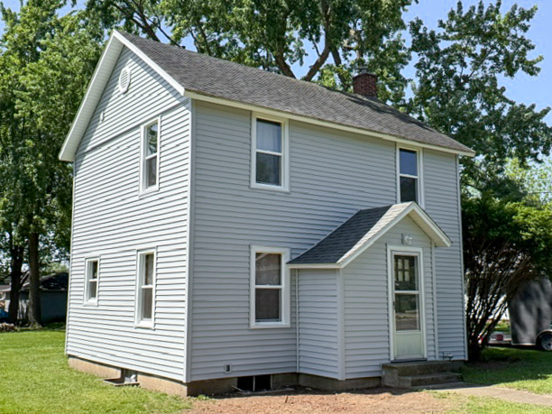 Two-story house with light gray siding and a small front porch, surrounded by grass and trees on a sunny day.