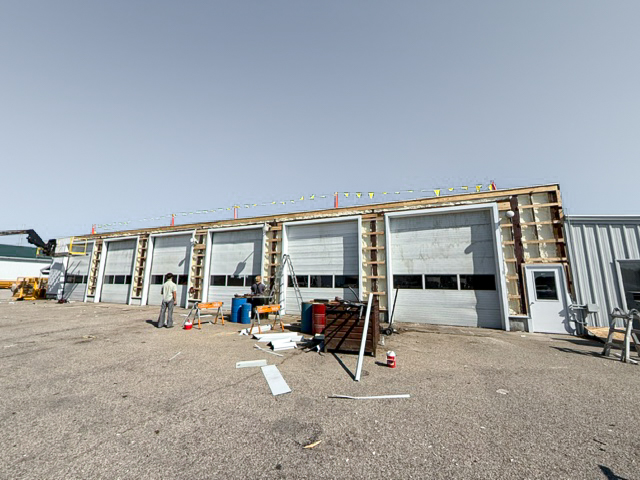 A commercial garage building under construction, with five large bay doors partially framed, tools and materials scattered outside, and workers present.