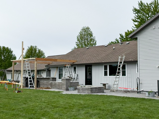 A backyard construction scene with ladders against a house, partially built patio structures, and building materials on the lawn.