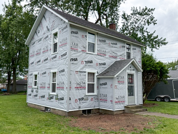Two-story house with Typar house wrap covering exterior walls, partially completed siding, and a small porch at the front, surrounded by grass and trees.