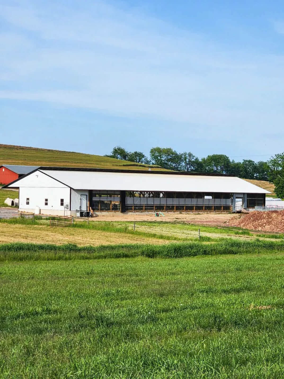 PREMEX Metal Roofing, Siding, & Trim Mifflinburg, PA A large barn with open sides sits on grassy farmland under a partly cloudy sky, with trees and another building in the background.