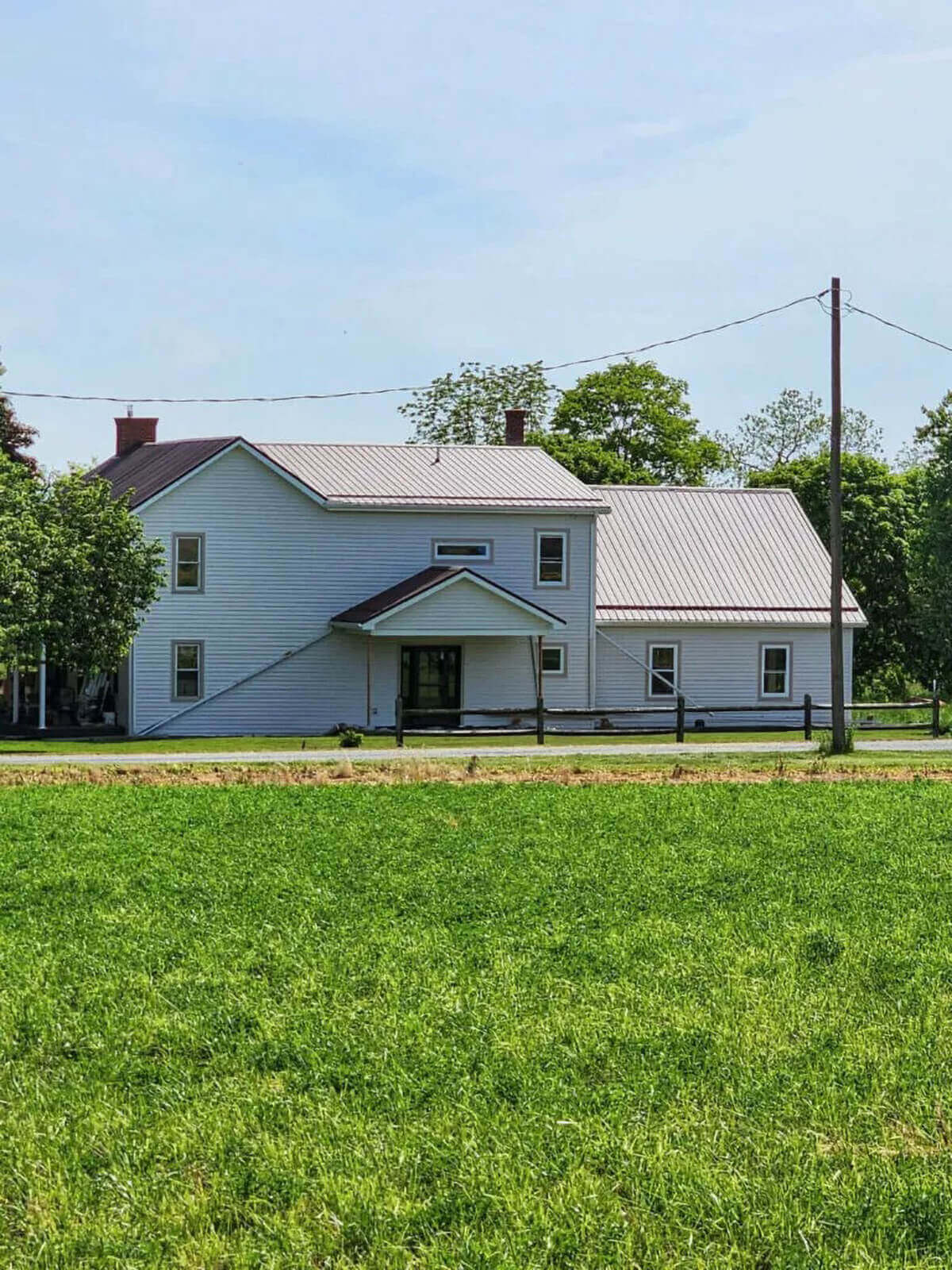 PREMEX Metal Roofing, Siding, & Trim Mifflinburg, PA A white two-story farmhouse with a metal roof sits behind a grassy field, bordered by trees and utility poles under a clear sky.