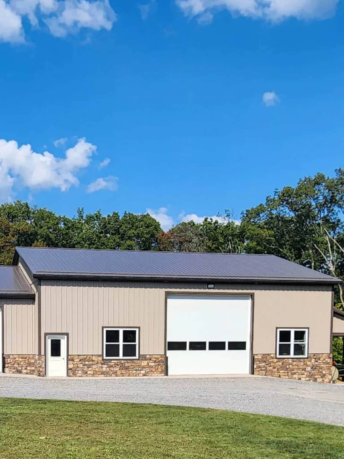 PREMEX Metal Roofing, Siding, & Trim Mifflinburg, PA A beige metal building with a stone base, large white garage door, small entry door, and windows sits on a gravel driveway with grass and trees in the background.