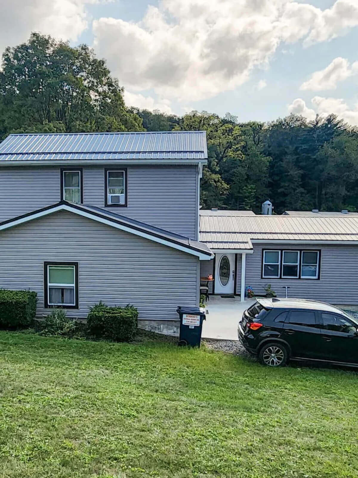 PREMEX Metal Roofing, Siding, & Trim Mifflinburg, PA A gray two-story house with a metal roof, a black car parked in the driveway, and green trees and grass surrounding the property.