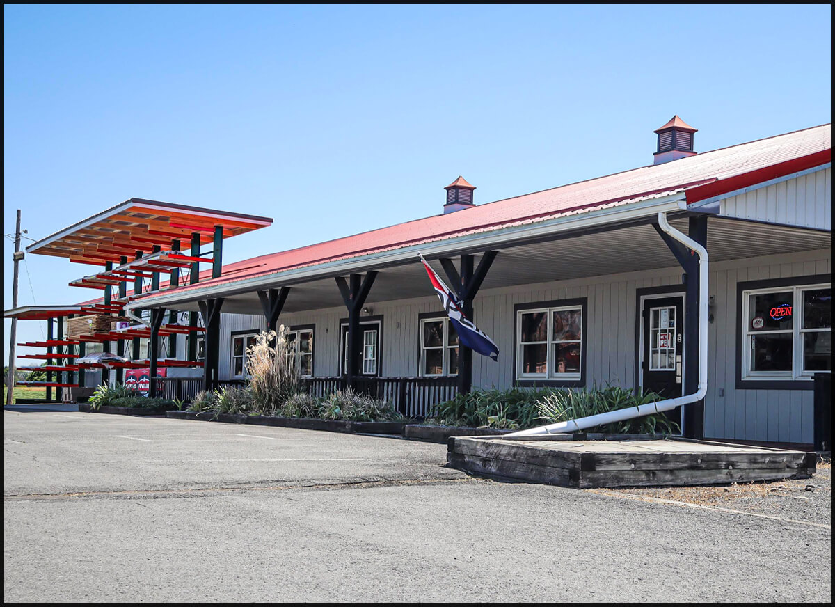 A metal building with a red roof, multiple windows, and an "open" sign. An American flag hangs by the entrance, and red metal shelves stand to the left. The parking lot is empty.