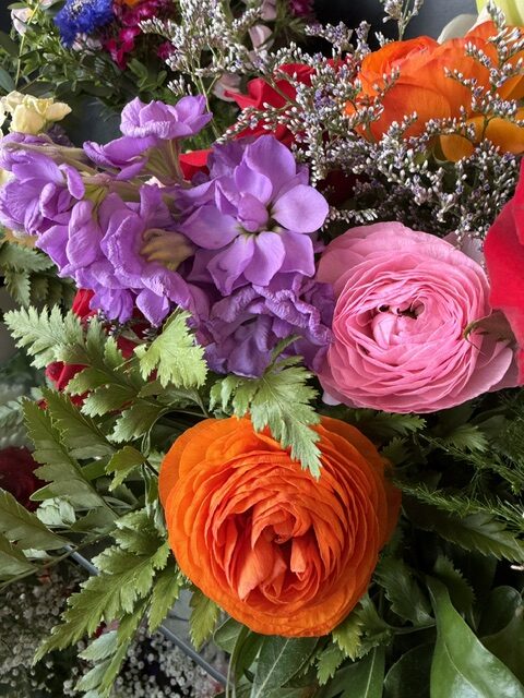 Swampy Hollow Greenhouse Gallery Cochranville, PA A close-up of a floral arrangement with orange, pink, and purple flowers surrounded by green leaves and small white blossoms.