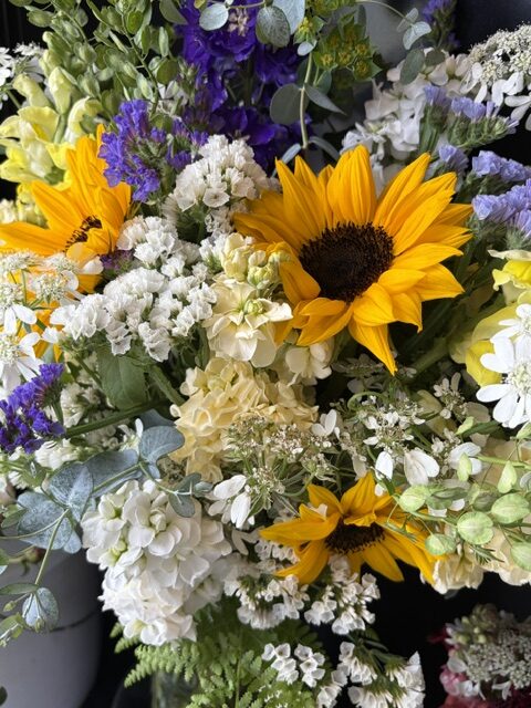 Swampy Hollow Greenhouse Gallery Cochranville, PA A bouquet featuring large yellow sunflowers, white hydrangeas, and small purple and white flowers with green foliage.