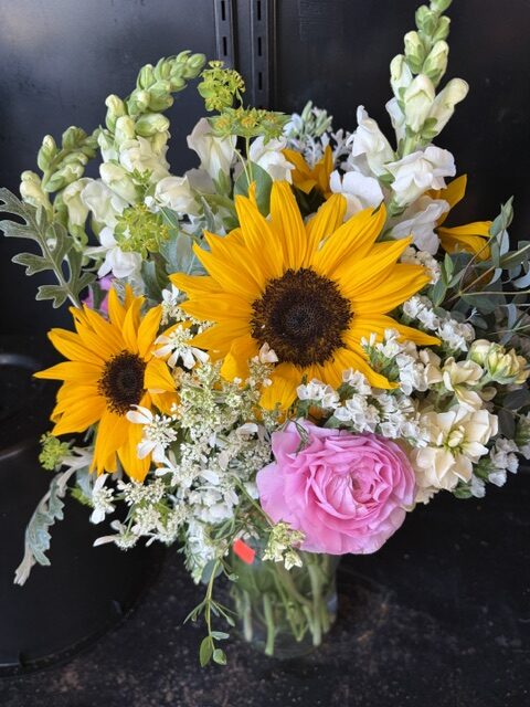 Swampy Hollow Greenhouse Gallery Cochranville, PA A bouquet with bright yellow sunflowers, white snapdragons, small white filler flowers, and a large pink rose arranged in a glass vase.
