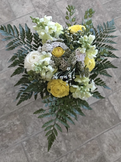 6.8.26 Swampy Hollow Greenhouse Bouquets Gallery 8 A bouquet of yellow and white flowers with green ferns, arranged in a circular pattern, placed on a tiled floor.