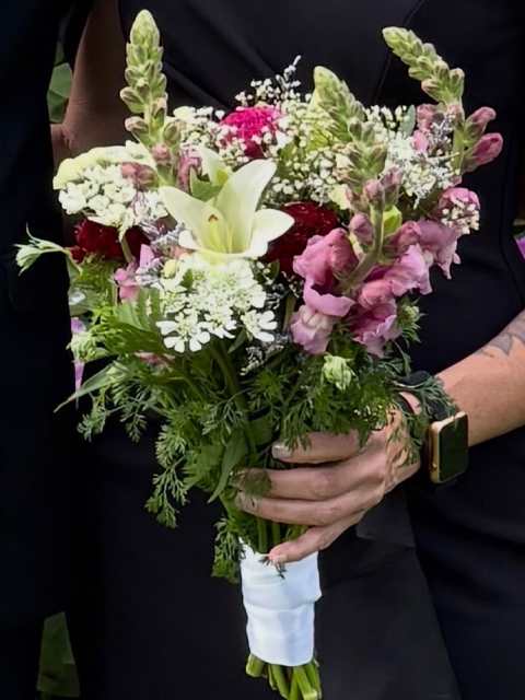 Swampy Hollow Greenhouse Gallery Cochranville, PA A person wearing a black outfit and smart watch holds a bouquet of mixed flowers, including lilies, snapdragons, and baby's breath, wrapped with white ribbon.
