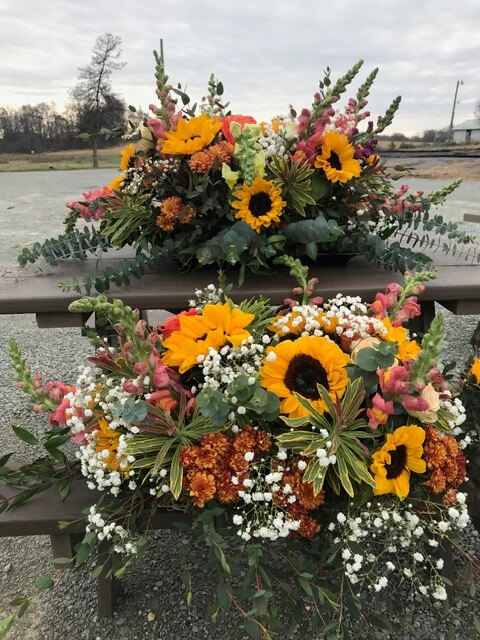 Swampy Hollow Greenhouse Gallery Cochranville, PA Two wooden benches display floral arrangements with sunflowers, snapdragons, baby's breath, and various greenery outdoors under a cloudy sky.