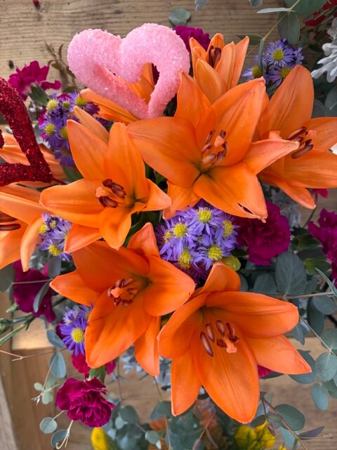 Swampy Hollow Greenhouse Gallery Cochranville, PA A bouquet featuring orange lilies, purple and yellow flowers, red flowers, greenery, and two pink and red heart decorations.