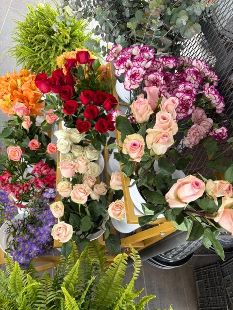 Swampy Hollow Greenhouse Gallery Cochranville, PA Various bouquets of roses and other flowers in different colors are arranged in buckets on display, surrounded by green plants.