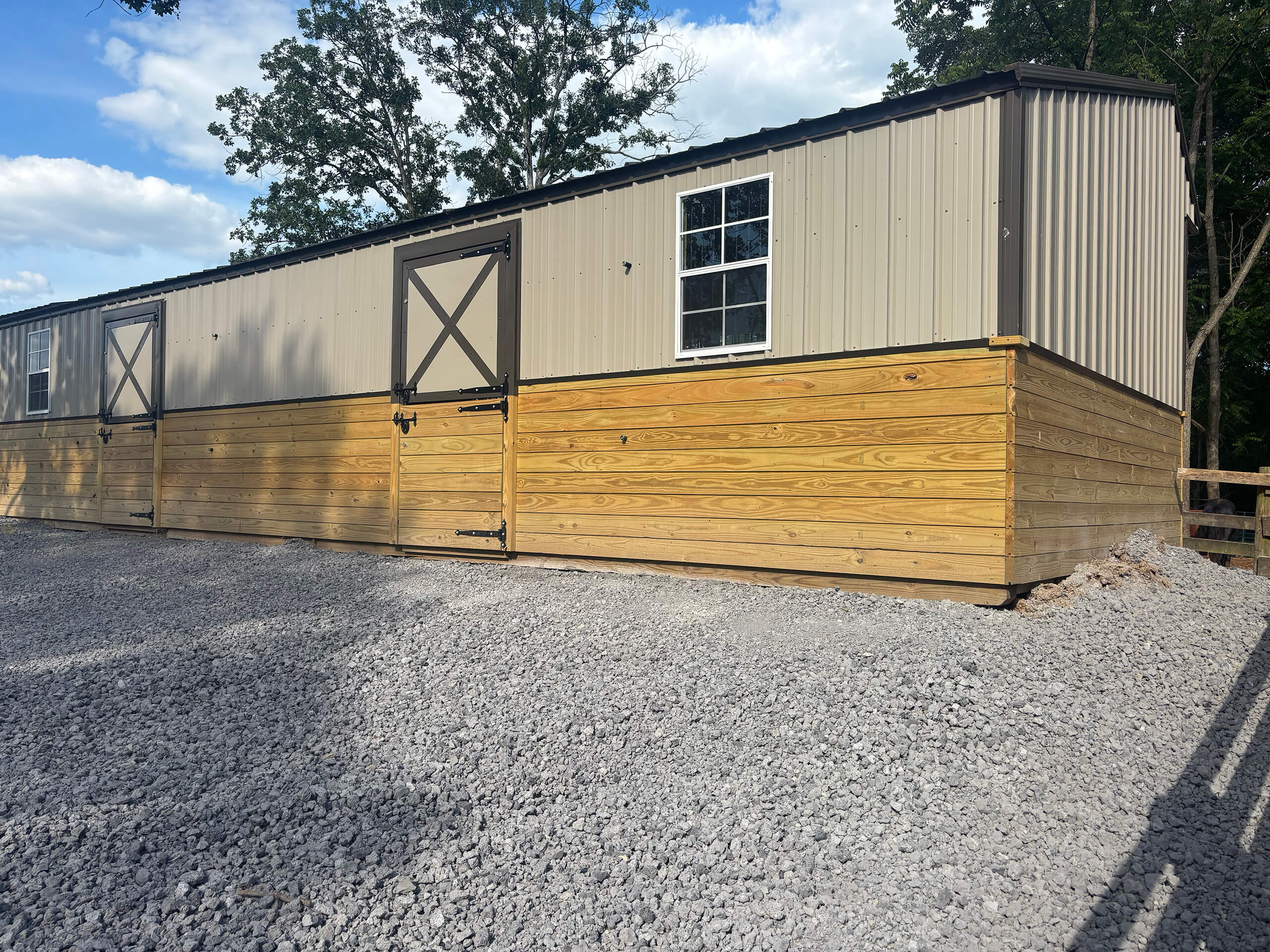 Frontier Land Services Excavating Equipment Irvington, KY A long, tan and brown metal and wood building with two barn-style doors and a window sits on a gravel lot under a partly cloudy sky.
