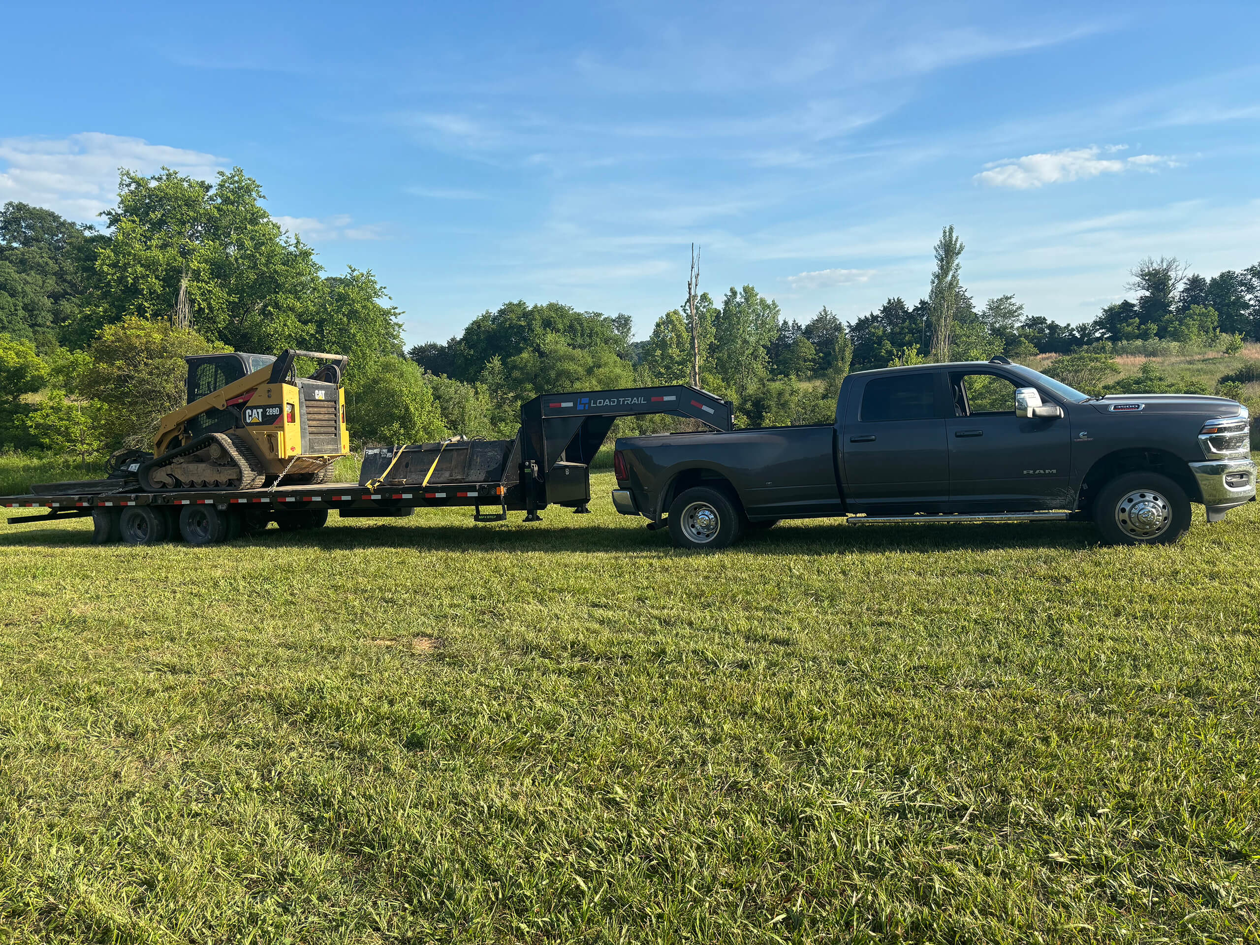 Frontier Land Services Excavating Equipment Irvington, KY A black pickup truck with a flatbed trailer carrying a yellow skid steer loader is parked on a grassy field with trees in the background.