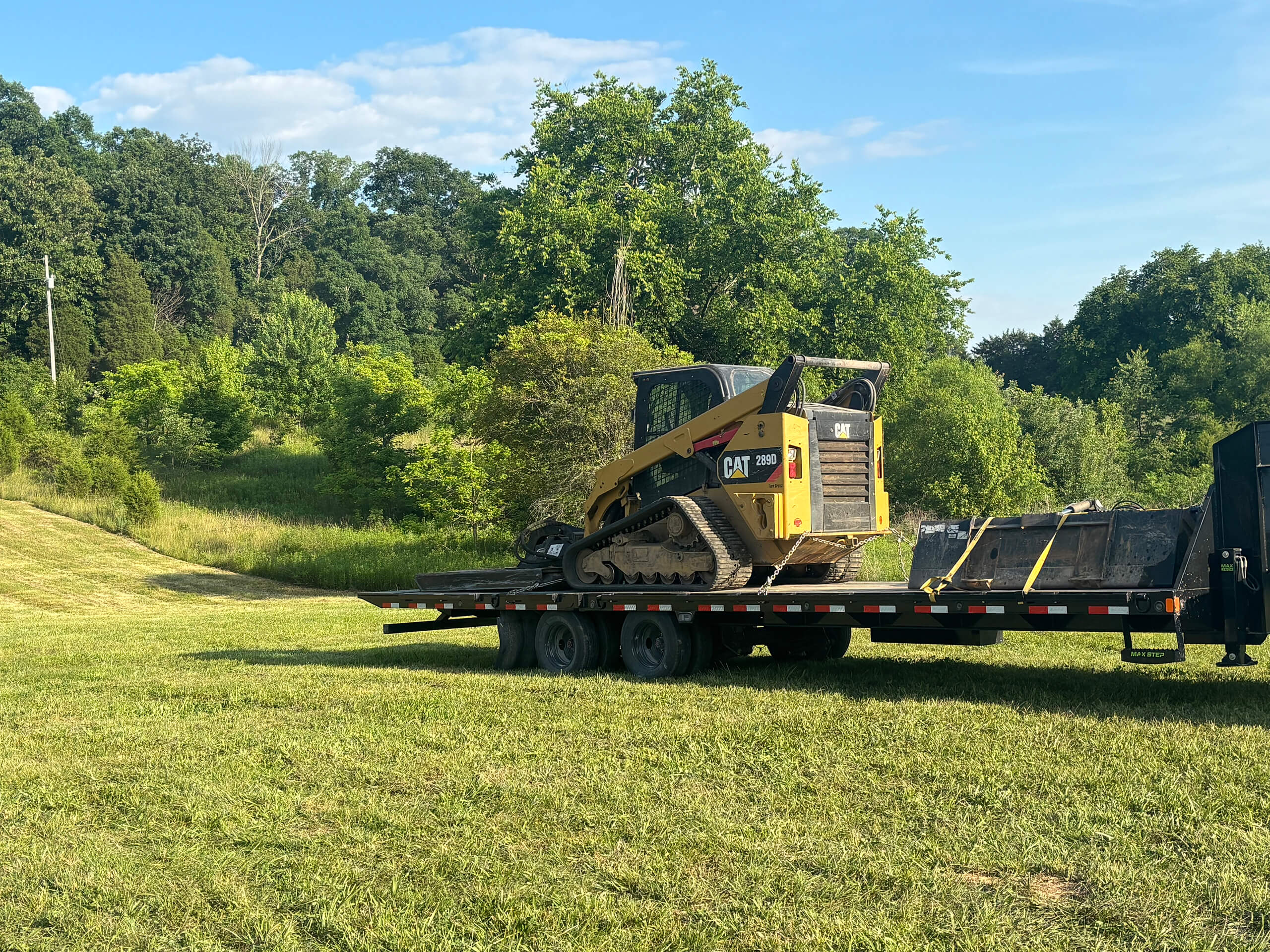 Frontier Land Services Excavating Equipment Irvington, KY A yellow Caterpillar skid steer loader is parked on a flatbed trailer in a grassy, tree-lined rural area under a partly cloudy sky.