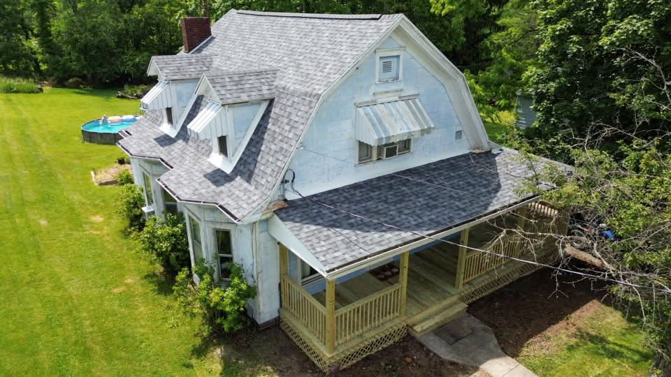A white, two-story house with a large covered front porch, grey shingled roof, and surrounding green lawn, with trees and a round pool in the background.