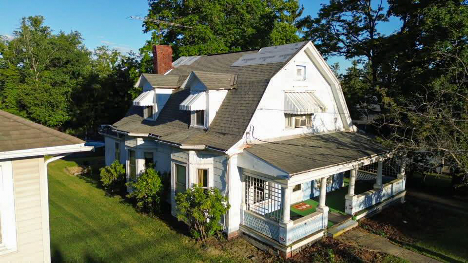 A two-story white house with a gray roof, front porch, overgrown shrubs, and visible signs of exterior wear, surrounded by trees and grass on a sunny day.