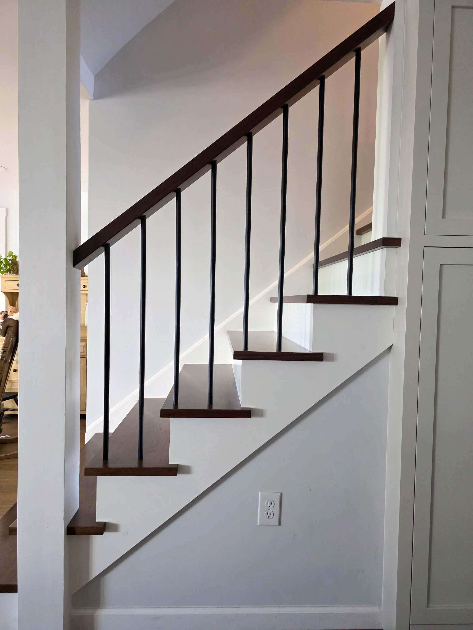 Brookview Builders Penn Yan, NY A staircase with dark wooden steps and black vertical railings runs alongside a white wall with a power outlet underneath.