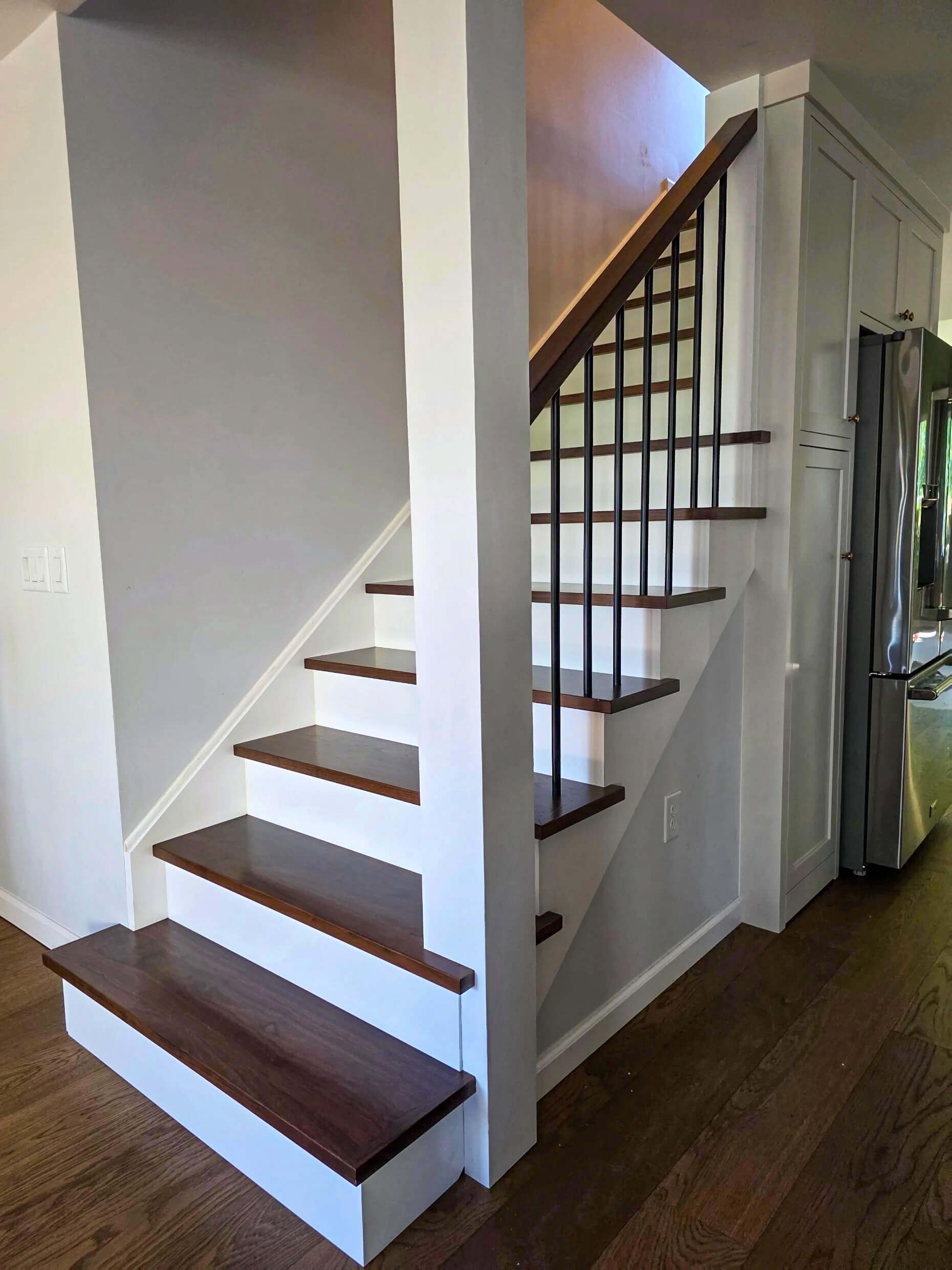 Brookview Builders Penn Yan, NY A staircase with dark wooden steps, white risers, and a black railing is shown next to a kitchen with a stainless steel refrigerator.