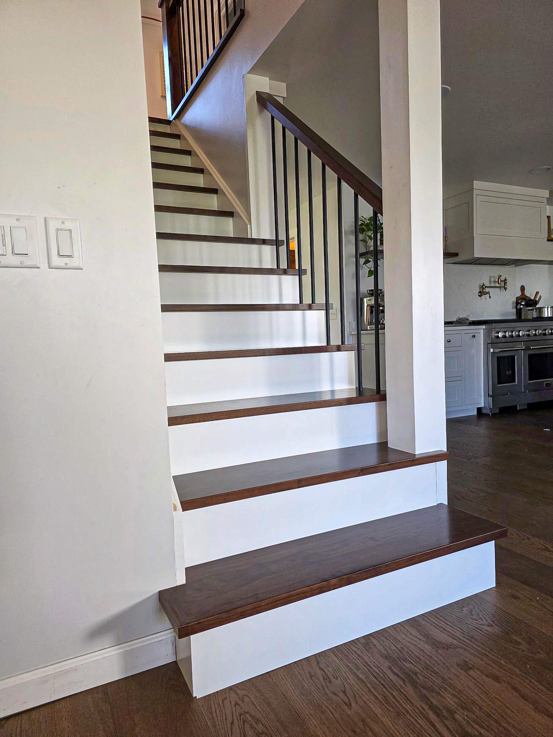 Brookview Builders Penn Yan, NY A wooden staircase with white risers and dark brown treads leads to an upper floor in a modern home with a kitchen visible in the background.