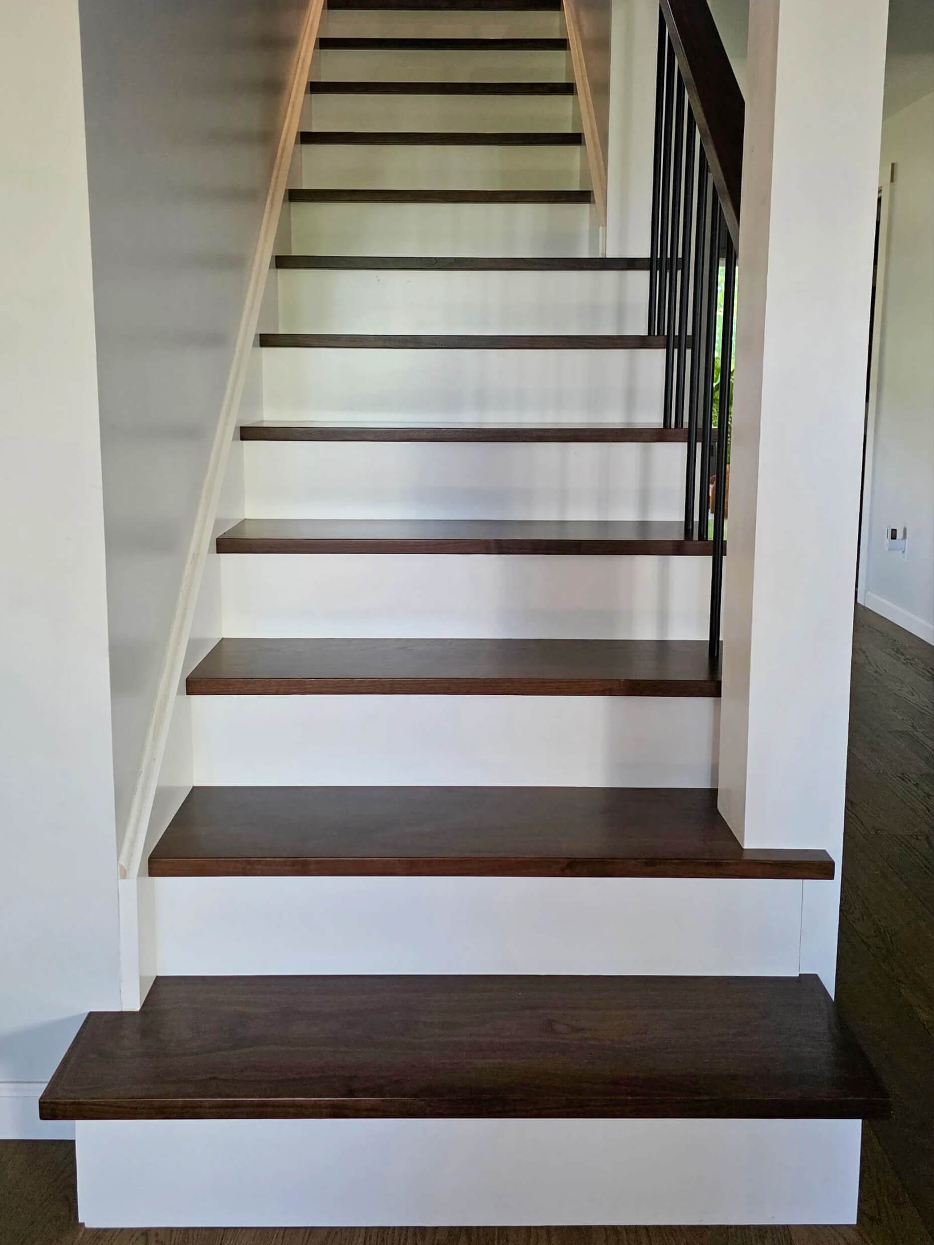Brookview Builders Penn Yan, NY A straight staircase with dark wooden treads and white risers, bordered by white walls and a black metal railing on the right side.