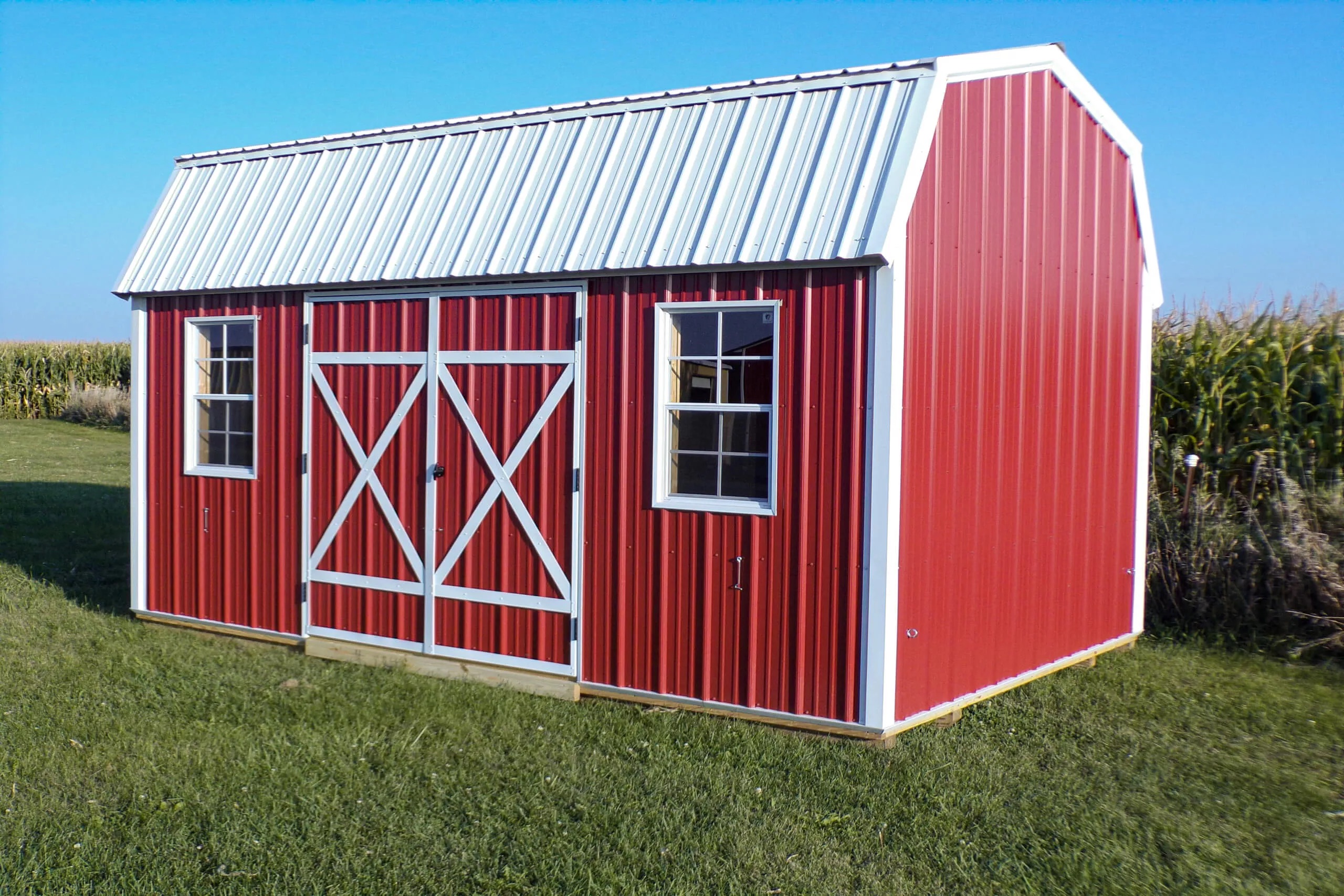 Red barn-style shed with white trim and a metal roof, featuring two windows and double doors, situated on a grassy area near a cornfield under a clear blue sky.
