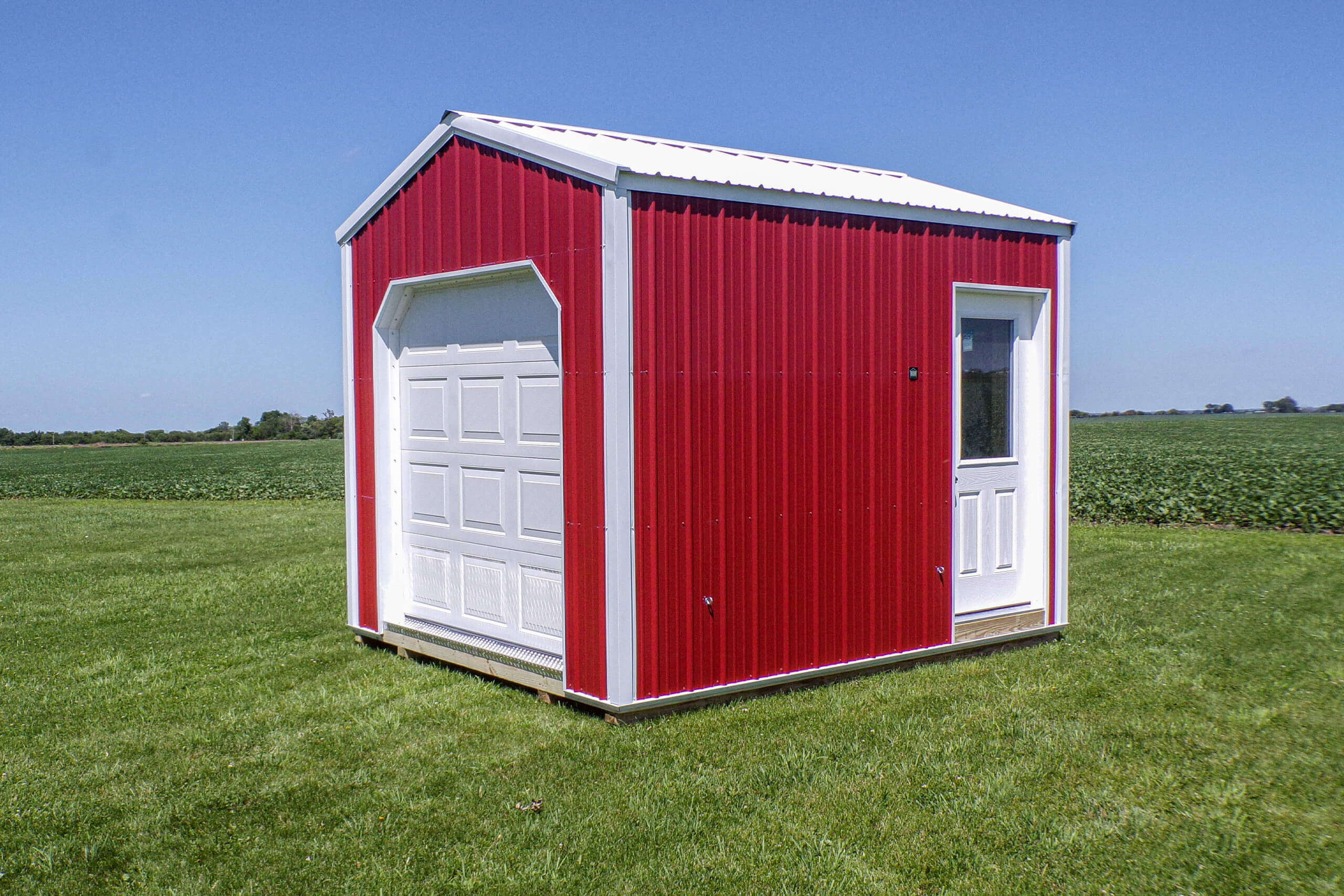 A small red metal shed with a white garage door and entry door sits on a grassy field under a clear blue sky.
