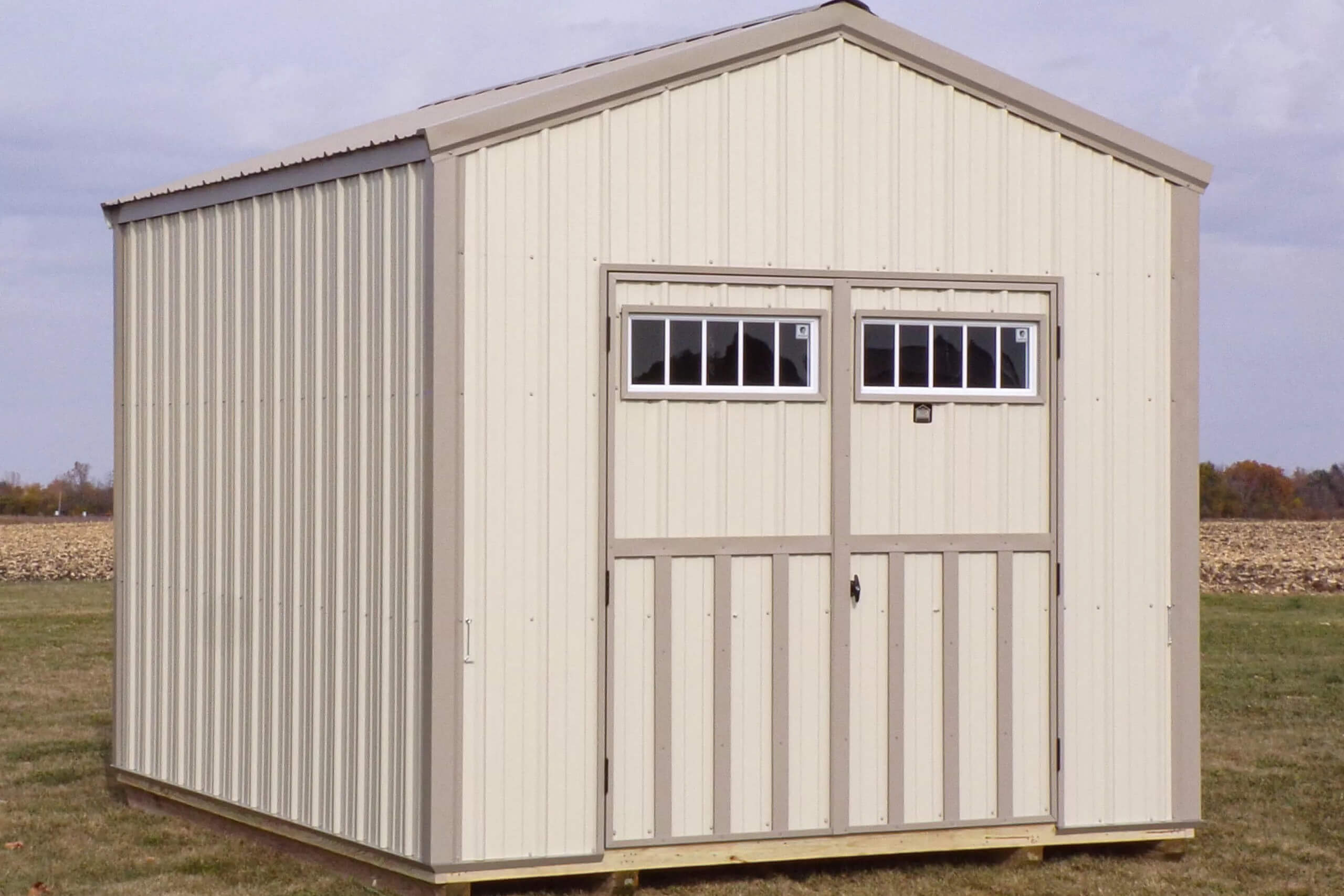 A beige metal storage shed with double doors and small windows stands on a grassy area with open land in the background.