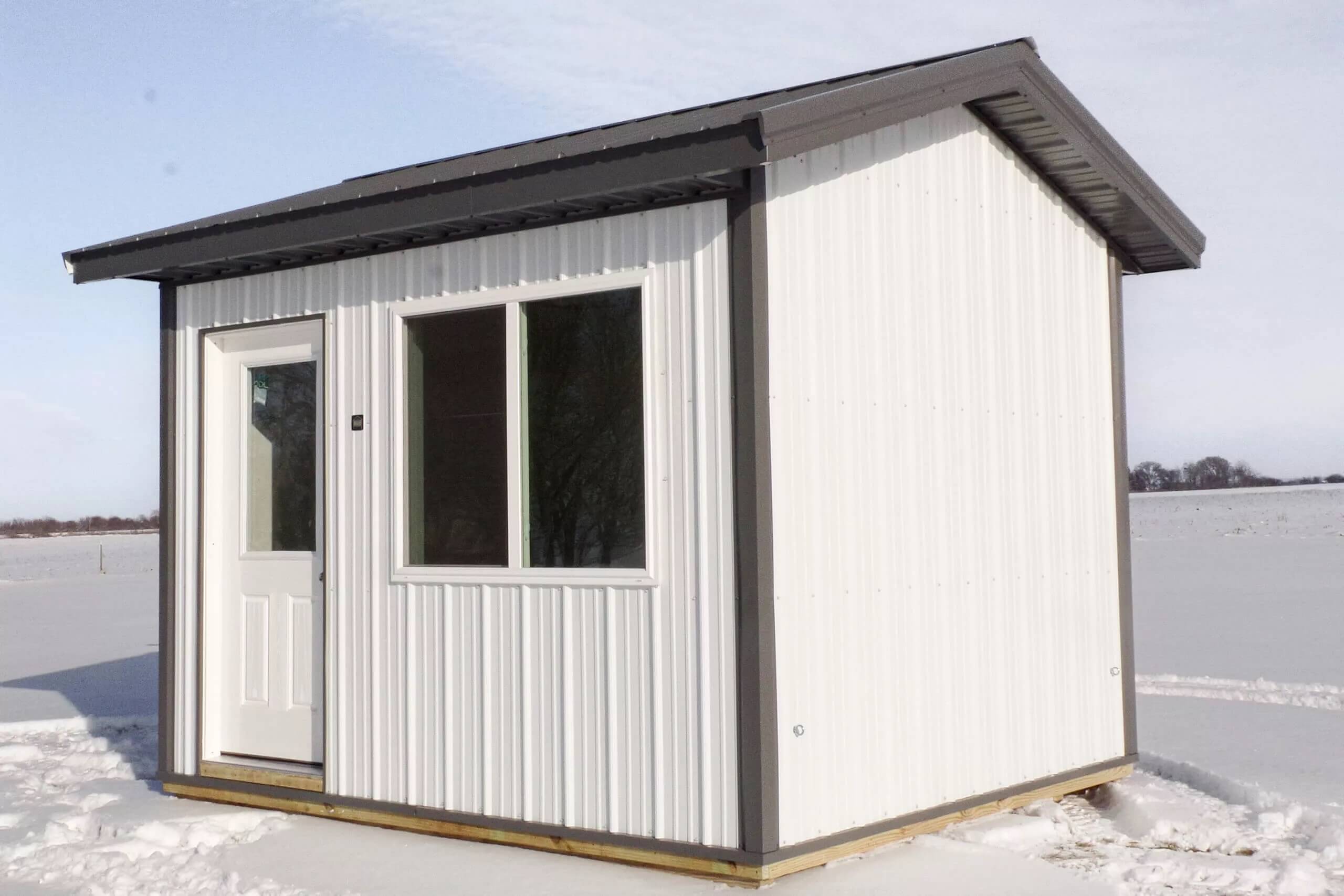 A small white metal shed with a door and two windows sits on a snowy field under a partly cloudy sky.